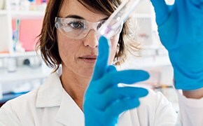 一位科学家身穿白大褂、戴着安全眼镜和蓝色手套，正在实验室里仔细检查一支装有粉红色溶液的试管，这说明研究工作涉及聚合物微球和纳米颗粒。
