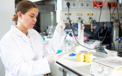 A laboratory technician in a white lab coat is preparing samples for food testing. The technician is holding a pipette and working at a lab bench.