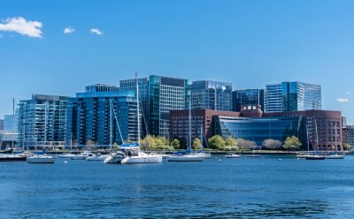 A scenic view of Boston's seaport skyline along a waterfront, featuring several high-rise buildings with glass facades and a mix of architectural styles. In the foreground, small boats are anchored in the water, and the sky is clear with a few clouds.