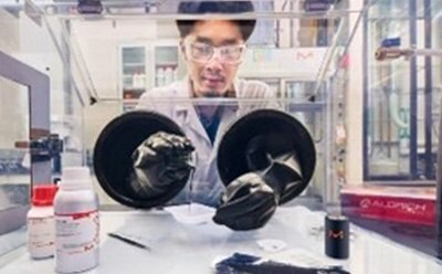 A scientist wearing safety goggles and a lab coat is working with a glove box in a high-tech laboratory. The image is taken from the outside of the glove box, showing the researcher through the transparent front panel. Various lab supplies, including chemical bottles and materials, are arranged on the bench in the foreground. The laboratory has a modern, industrial setting with advanced equipment in the background. A scientist wearing safety goggles and a lab coat is working with a glove box in a high-tech laboratory. The image is taken from the outside of the glove box, showing the researcher through the transparent front panel. Various lab supplies, including chemical bottles and materials, are arranged on the bench in the foreground. The laboratory has a modern, industrial setting with advanced equipment in the background.