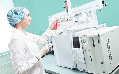 Female scientific researcher with lab coat, gloves and hair protection placing vial flask in gas chromatography instrument.