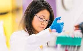 The image depicts a professional setting where a person is wearing a white lab coat and bright blue latex gloves. They are engaged in a precise task, handling equipment or conducting an experiment in a laboratory environment filled with various blurred apparatuses in the background.