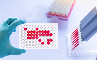 Laboratory setting where a researcher, wearing a blue glove, is holding a white microtiter plate with red wells. The plate is prominently labeled “ANIMAL COMPONENT FREE,” indicating the use of animal-free reagents in the experiment. In the background, there are racks filled with pipette tips, highlighting the precision and cleanliness of the scientific work being conducted. 