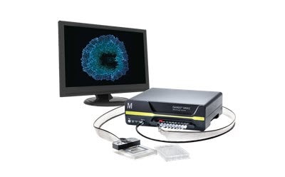 A computer monitor connected to a black electronic device with multiple ports, showcasing a blue circular pattern on the screen. Two small rectangular objects are placed in front of the device, and two cables connect the monitor to the device. 