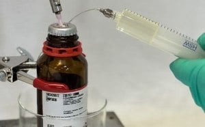 A close-up image of a laboratory setting showing a brown glass bottle of organometallic reagents with a white label and a Sure/Seal™ cap. A syringe is being used to extract liquid from the bottle, with a thin needle supplying inert gas inserted through the cap. The person handling the syringe is wearing a green glove, indicating safety precautions. The background is blurred, focusing on the bottle and syringe, with a glass container visible beneath the bottle.