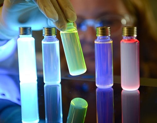 The image depicts light-emitting materials for organic light emitting diode (OLED).  A person wearing gloves is holding a glowing green vial among other brightly colored glowing vials on a reflective surface. The vials emit various colors, including pale blue, yellowish-green, deep purple, and bright red. The person’s face and protective goggles are subtly outlined by the glow, and each vial has a metallic cap. The reflection of the glowing vials is visible on the surface. 