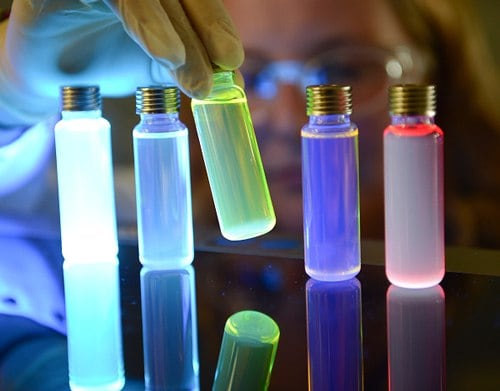 Vials of OLED materials lit up by UV light The image depicts light-emitting materials for organic light emitting diode (OLED). A person wearing gloves is holding a glowing green vial among other brightly colored glowing vials on a reflective surface. The vials emit various colors, including pale blue, yellowish-green, deep purple, and bright red. The person’s face and protective goggles are subtly outlined by the glow, and each vial has a metallic cap. The reflection of the glowing vials is visible on the surface.