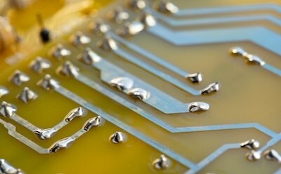 A close-up of an electronic circuit board featuring conductor traces. The traces are highlighted by shiny silver drops of solder, set against a vibrant yellow background.