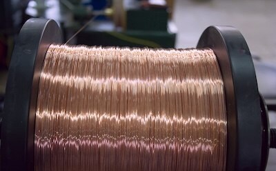 A close-up of a reel of copper wire in a manufacturing setting. The wire is neatly coiled, showcasing its shiny, reflective surface.