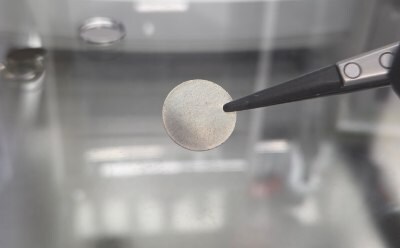 A metallic disc held by tweezers, set against a blurred background.