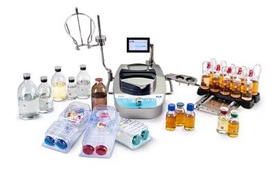 An assortment of sterility testing consumables, media, and instruments. At the center, there’s a sterility testing instrument with a digital screen and attached tubing. Surrounding the instrument are multiple bottles of different sizes containing clear and amber liquids. There are also several petri dishes with colored media in blue, red, and clear hues. To the right side of the image, there’s a rack holding vials with red caps, possibly containing samples or culture media. The background is white.