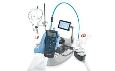 A Steritest Symbio pump system used for sterility testing in a laboratory setting. The main focus is a handheld device with a digital screen, and several buttons below the screen. This device is connected by a blue tube to the main pump system, which has various ports. In the bbackground, there are various laboratory items. The emphasis on the handheld device suggests it’s an important part of maintenance or monitoring the Steritest Symbio system.