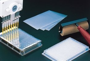 A multi-channel pipette dispensing liquid into a well plate, with AlumaSeal 96 ® foil, a stack of filter papers, and a roller bottle nearby, all placed on a dark surface against a black background.