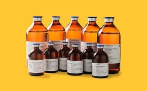 A collection of amber glass bottles with white labels, containing cell separation media such as Ficoll-Paque<sup>®</sup> and Percoll<sup>®</sup>, against a yellow background. Several amber glass bottles of varying sizes are arranged in two rows. The bottles have white labels with black text.