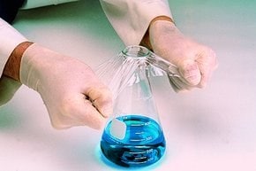 A person wearing lab gloves is applying a sealing film to a flask containing a blue liquid