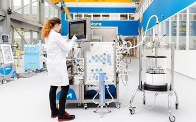 A laboratory setting showcasing the tools and chemicals used in affinity chromatography. In the foreground, three bottles labeled with hazard symbols indicate the presence of flammable and corrosive substances, essential for selective biochemical interactions and purification processes. In the background, a scientist in a lab coat is engaged with complex equipment, likely related to large-scale manufacturing and process development in affinity chromatography.