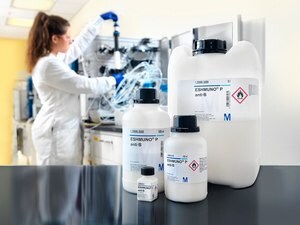 A scientist in a lab coat working with equipment and chemicals in a laboratory setting. In the foreground, there are three chemical containers with labels, two are large and one is smaller, marked with hazard symbols for flammable and corrosive substances.