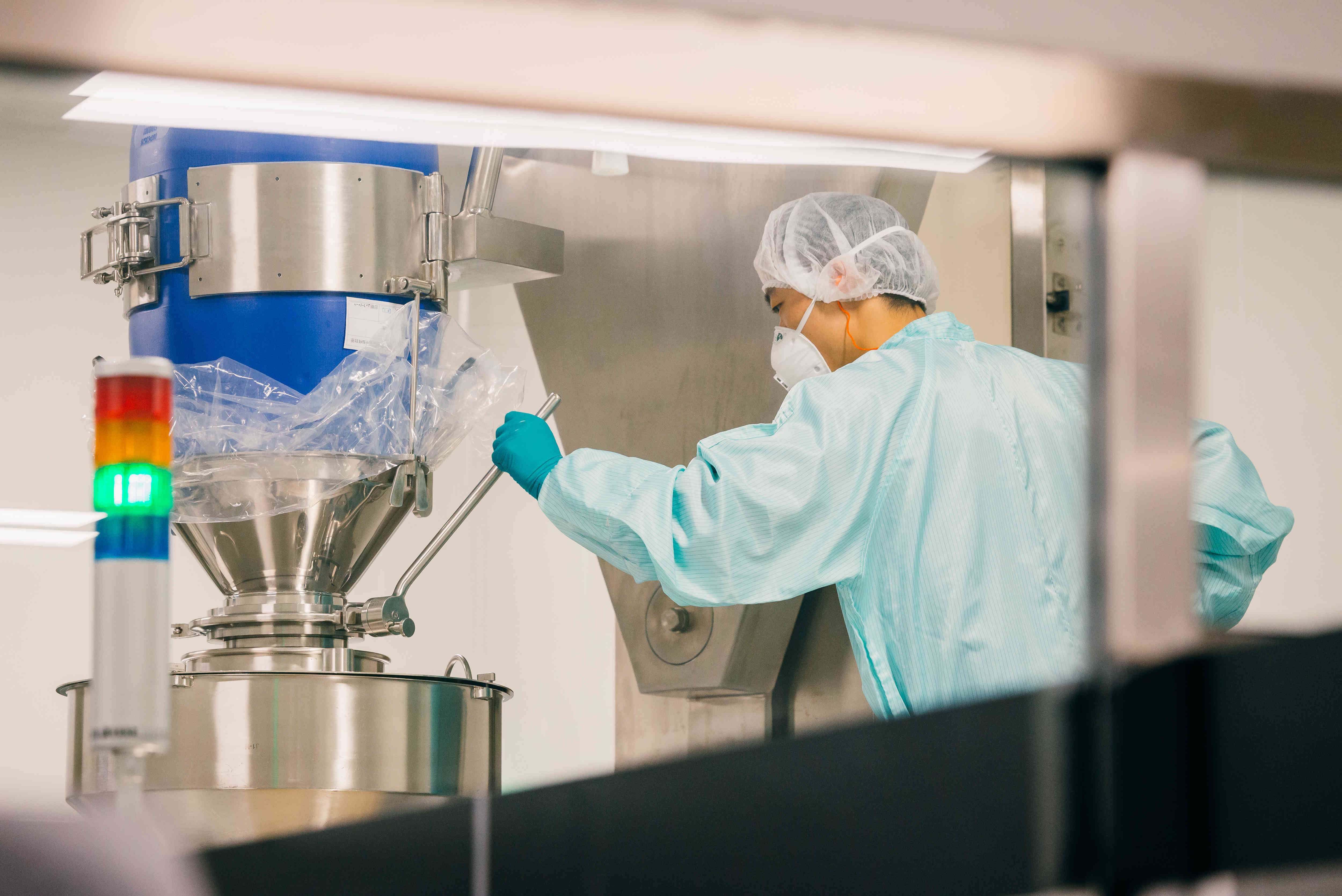 The image depicts a scientist in protective gear, who is transferring materials from a plastic bag into a stainless steel funnel connected to a metallic machine in a sterile environment, possibly for pharmaceutical or food processing purposes.