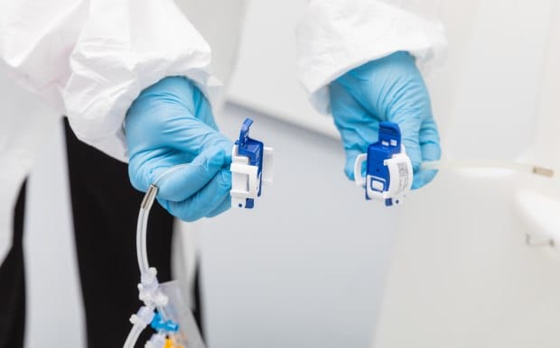 A person waring a lab coat and gloves holds a NovaSeptum® SURe sampling assembly that ends in a blue connector in one hand, and tubing of a single-use process  ending in a blue connector in the other, seemingly intending to connect them.