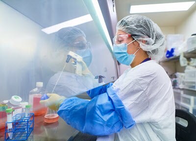 The image depicts a person in a laboratory setting, engaged in scientific work. They are wearing a white lab coat, blue gloves, and a hairnet, which indicates adherence to safety protocols. The individual is handling test tubes containing colored liquids, and various lab equipment and chemicals are arranged on the counter beside them. The background shows shelves stocked with additional lab supplies and equipment, emphasizing the professional and well-equipped nature of the laboratory.