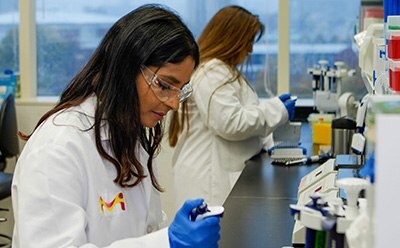 一位科学家坐在实验台前，戴着实验室护目镜、白大褂和手套。她正在移液。在她身后，另一位身穿白大褂、戴着护目镜的科学家正在使用多通道移液器。图片右侧是仪器和移液器吸头。