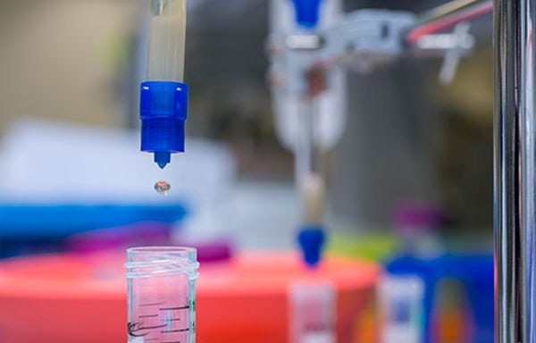 A close-up of a laboratory setting. The main focus is on a blue pipette dispensing a droplet into a small glass container partially filled with a pink-colored liquid. The background, although blurred, suggests the presence of various other laboratory instruments or containers.