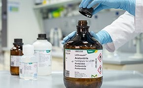 Large, brown solvent bottle with Supelco® branded label is opened by a lab worker wearing blue nitrile gloves. Only the hands and lower arms of the worker are shown. One small brown solvent bottle and two small white bottles can be seen in the background.