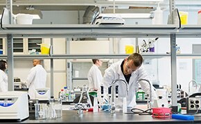 Five scientists wearing white lab coats, safety goggles and blue nitrile gloves work at different stations within the lab operating chemistry analytical instruments and filling glass beakers and vials. 