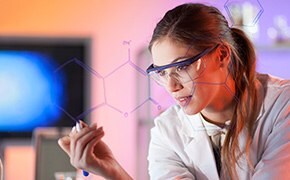 身穿白色实验服和护目镜的棕发女科学家在玻璃屏幕上书写化学结构。背景可见电脑屏幕和实验室环境。