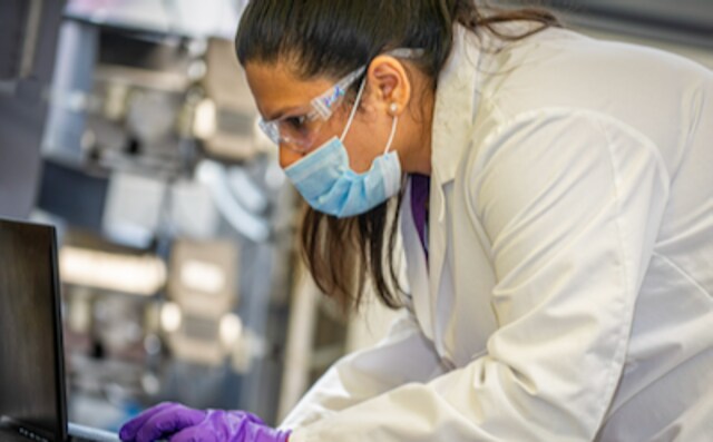 Researcher enters data in a computer in a lab setting, wearing glasses, mask, gloves and a lab coat