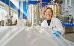 Shannon 正穿着实验室外套、戴着蓝色手套和护目镜，站在M Lab（非 GMP 生物制造环境）中。她正微笑地拿着一只垃圾袋（左下角）。