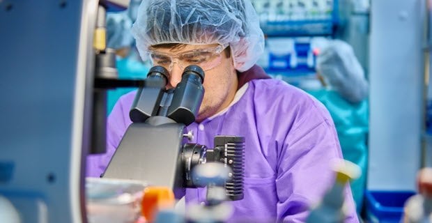 穿着紫色实验室外套、戴着护目镜和发套的科学家正在观察显微镜。