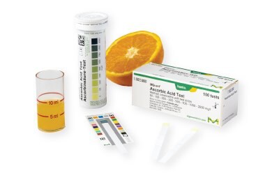 A citrus fruit and a beaker containing a liquid sample from the fruit which is subject to Ascorbic acid testing using MQuant® Ascorbic Acid Test Strips, alongside MQuant® Color Reference Card and packages of the test strips