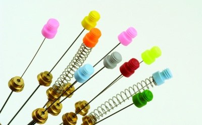 The image appears to be a collection of metallic colorful push pins and springs. The pin heads are of plastic and contain pink, green, yellow and other colors. 