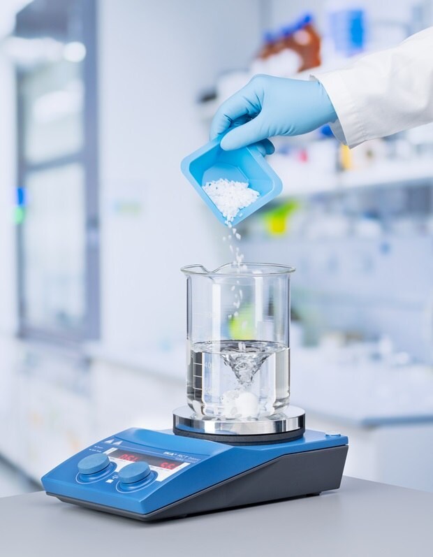 A gloved hand pours white granules from a blue container into a clear beaker with swirling water on a blue magnetic stirrer in a bright lab.