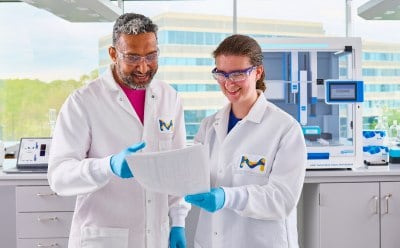 2 scientists standing in front of AAW™ automated assay workstation holding paper and chatting to each other.