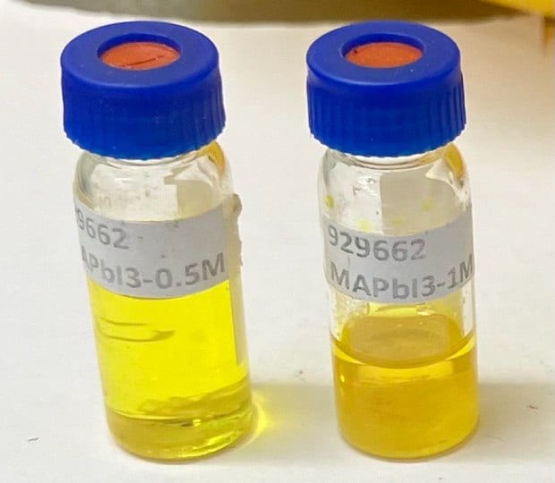 Two small glass vials with blue caps, placed next to each other on a white surface. The vial on the left is labeled '929662 MAPbI3-0.5 M' and contains a yellow solution, while the vial on the right is labeled '929662 MAPbI3-1.0 M' and features a deeper yellow color. The image illustrates the different concentrations of MAPbI3 dissolved in ElectroGreen® NMP Type-2 solvent, highlighting the impact of concentration on the solution's color.