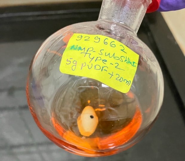 A round-bottom flask containing a bright orange solution, suspended in a laboratory setting. The flask is labeled with a bright yellow sticker that reads '929662 NMP Substitute Type-2 5g PVDF-20mL.' Inside the flask, a small white object, possibly a stirring bead or a sample, is visible. The image captures the preparation of a poly(vinylidene fluoride) (PVDF) solution using ElectroGreen® NMP Type-2 solvent, highlighting the color and contents of the mixture.