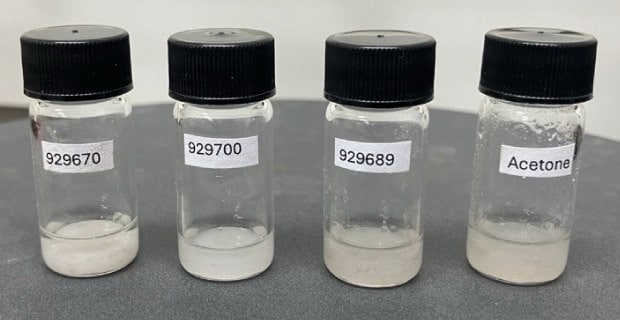 Four clear glass vials arranged side by side on a gray surface, each containing EPS dispersed in varying solvents. The vials are labeled with ElectroGreen® product numbers: 929670, 929700, 929689, and the last one labeled 'Acetone.' The contents of the first three vials appear as white or cloudy dispersions, while the vial containing acetone shows a clear liquid, indicating the different solvent properties and dispersions of the samples.