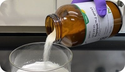 Amber reagent bottle labeled Supelpak 2 PFAS adsorbent pouring purified XAD-2 resin granules into a glass beaker for PFAS air emissions analysis.